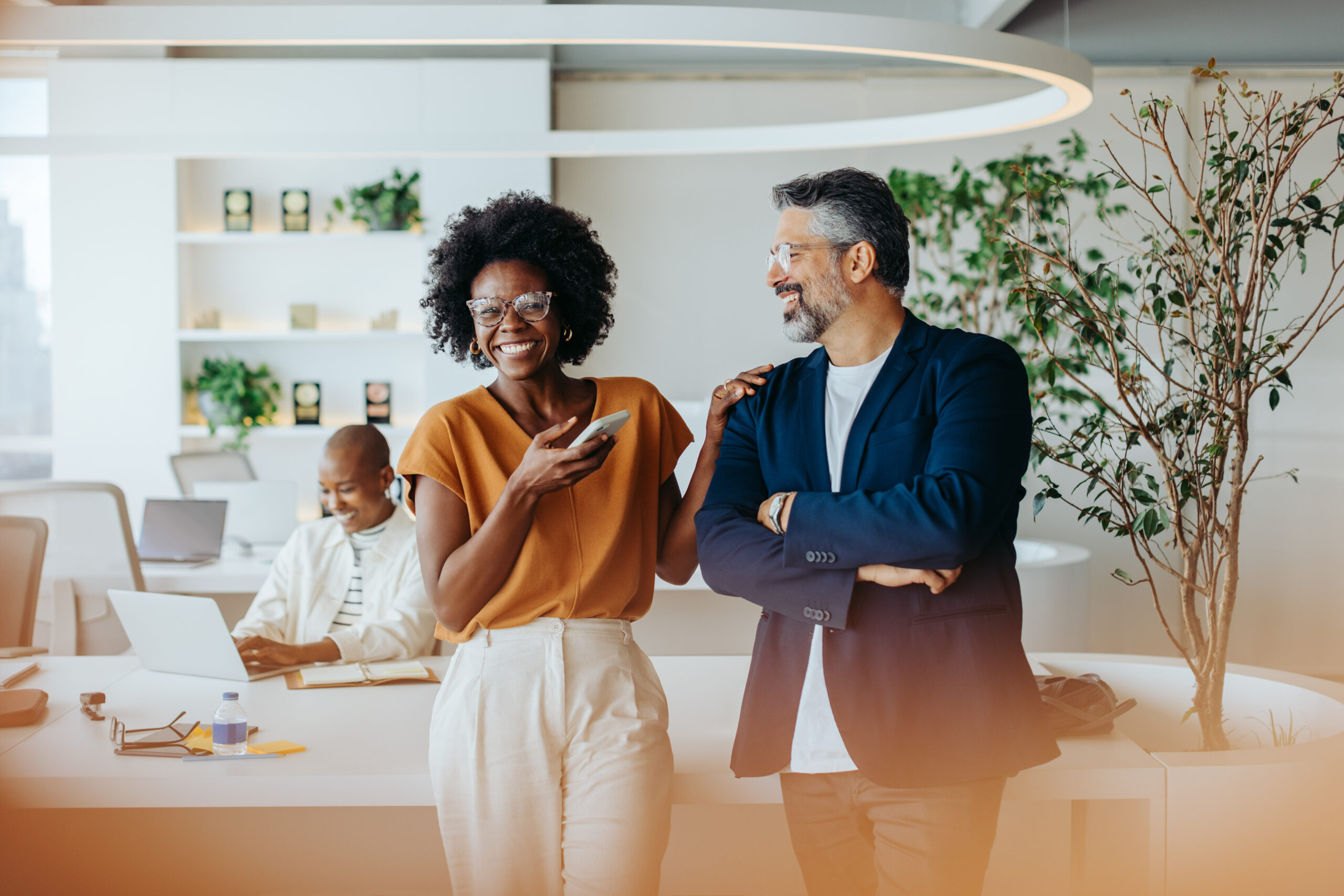 Two professionals working together in a modern open plan office. The colleagues, a business man and a business woman, are standing and smiling, demonstrating their teamwork and success.