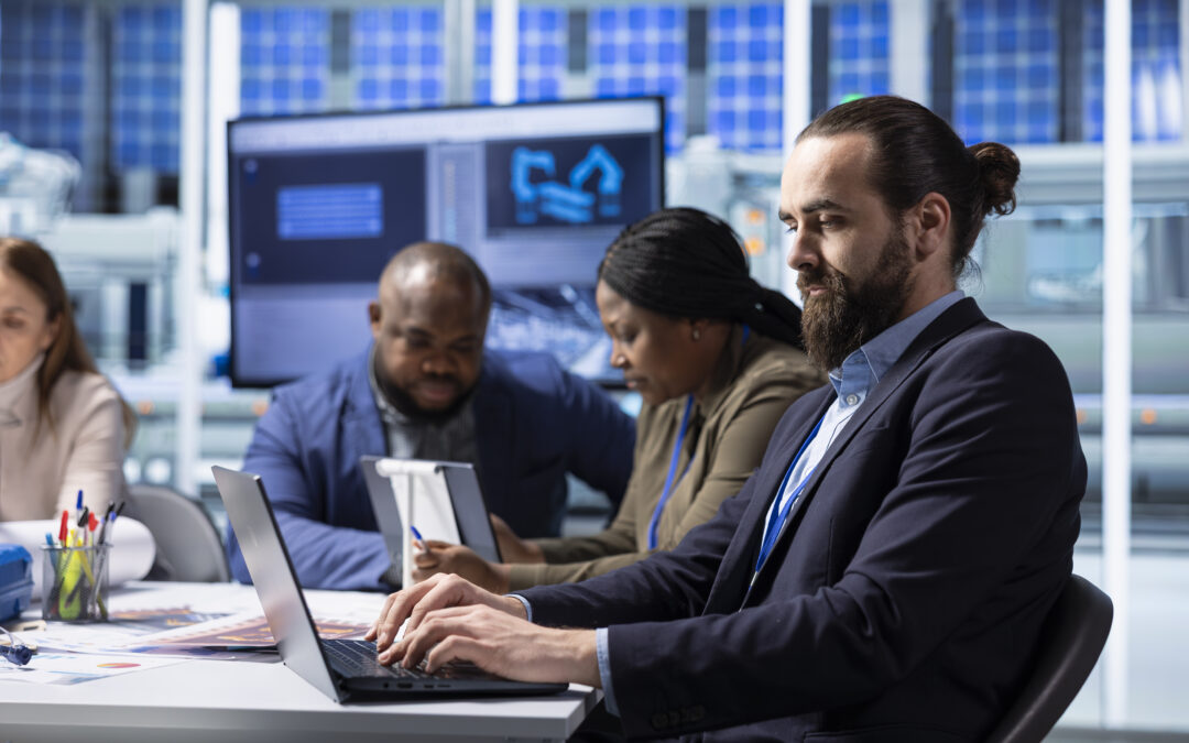 A diverse group of four professionals in business-casual attire working at a long white desk in a high-tech facility. In the foreground, a man with a beard and hair in a bun types on a laptop, while colleagues in the background review data on tablets and large monitor screens.