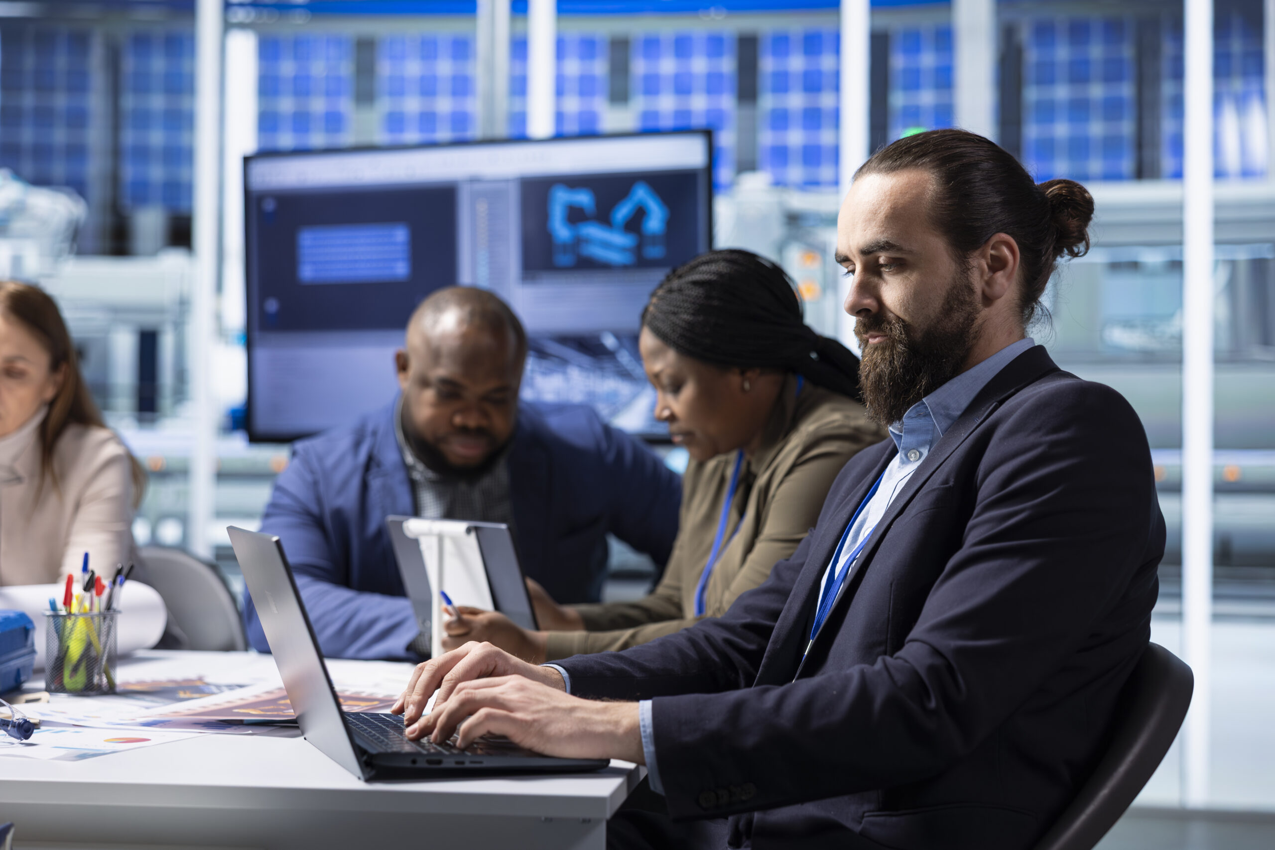 A diverse group of four professionals in business-casual attire working at a long white desk in a high-tech facility. In the foreground, a man with a beard and hair in a bun types on a laptop, while colleagues in the background review data on tablets and large monitor screens.